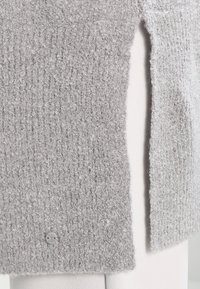 Grå texturerad tröja med ett ribbat mönster, sidokilar och en klar plastknappdetalj. Mjuk, stickad material med en mjuk yta.