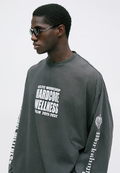 Muž so šedými slnečnými okuliarmi a voľnou šedou košeľou s dlhým rukávom, na ktorej je biely nápis "032C Workshop Hardcore Wellness Berlín 2025-2032."