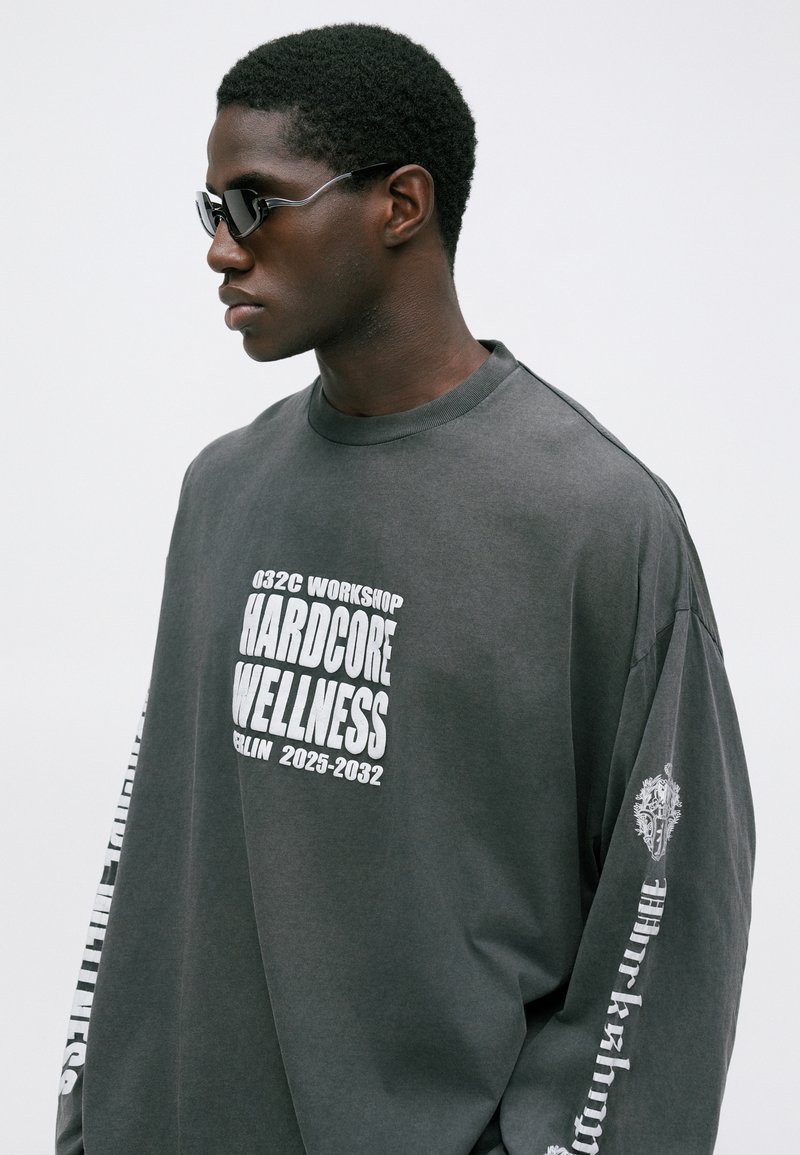 Mann mit dunklen Sonnenbrillen und einem lockeren grauen Langarmshirt mit weißem Text "032C Workshop Hardcore Wellness Berlin 2025-2032."