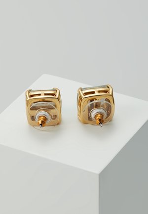 Złote kwadratowe kolczyki z zapięciem przezroczystym, prezentowane na białym piedestale na tle jednolitego jasnoszarego tła.