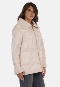 Giacca zip-up color crema realizzata in tessuto morbido, con un ampio colletto, due tasche frontali e cuciture sottili per la struttura.
