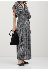 Femme portant une robe maxi noire et blanche à motif feuilles avec une bordure noire, tenant un sac à main noir tressé et portant des sandales slides noires.