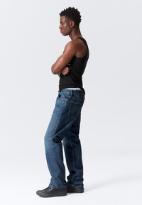 Un maieu negru și blugi bleu purtați de o persoană care stă de profil. Blugii au o croială lejeră, iar ținuta este completată cu pantofi negri.