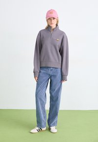 Hanorac gri cu fermoar, cu logo brodat, asortat cu blugi largi, decolorati, de culoare albastră și adidași albi cu dungi negre. Se poartă o șapcă roz.