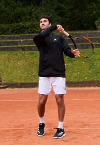 Mann i svart jakke og hvite shorts svinger en tennisracket på en leirgulv med grønne omgivelser og steintrapper i bakgrunnen.