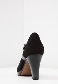 Chaussure à talon haut en daim noir avec un bout arrondi, une bride en cuir verni noir brillant et un détail de boucle, et un talon effilé.