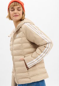 Mujer con chaqueta de plumas beige de Adidas con franjas blancas en las mangas, gorro de punto naranja y vaqueros azules, mirando de lado con la mano en el bolsillo.