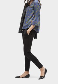 Blouse fleurie bleue à manches évasées, pantalon noir ajusté, et ballerines pointues noires. Veste en cuir portée sur une épaule.