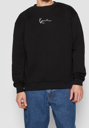 Man wearing black sweatshirt with small white cursive logo on chest and blue jeans, standing against plain light background.