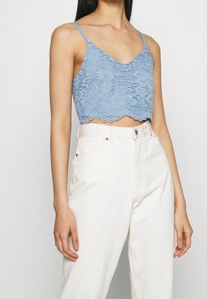 Frau trägt ein hellblaues, florales Spitzen-Cropped-Camisole mit Spaghetti-Trägern und hochtaillierte cremeweiße Hose.