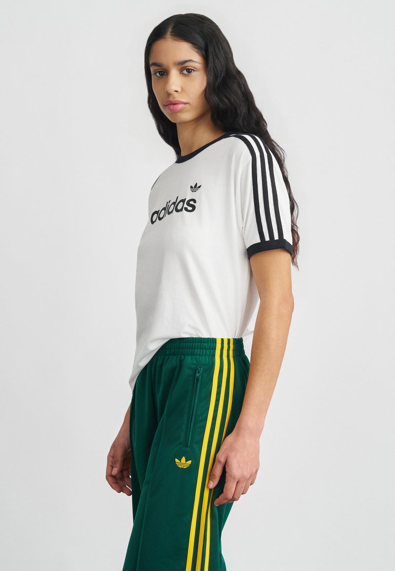White t-shirt with black ringer collar and three black stripes on sleeves, paired with green pants featuring yellow stripes and zip pockets.