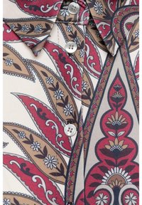 Een button-up shirt met een crèmekleurige basis en een gedurfd rood, bruin en zwart paisleypatroon, kleine witte bloemaccenten en zilveren knopen.
