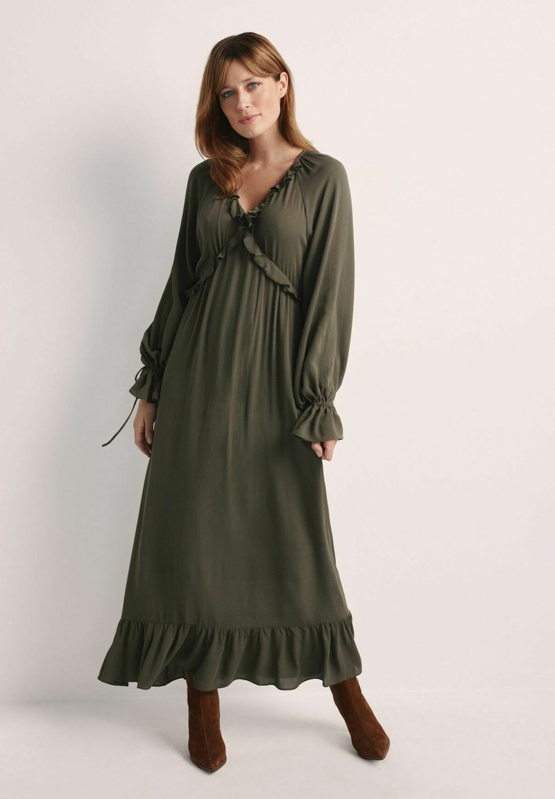 Oliwkowa sukienka maxi z falbaniastym dekoltem i rękawami, z p�łynącą sylwetką i falbaną u dołu. Nosi ją w połączeniu z brązowymi botkami za kostkę.
