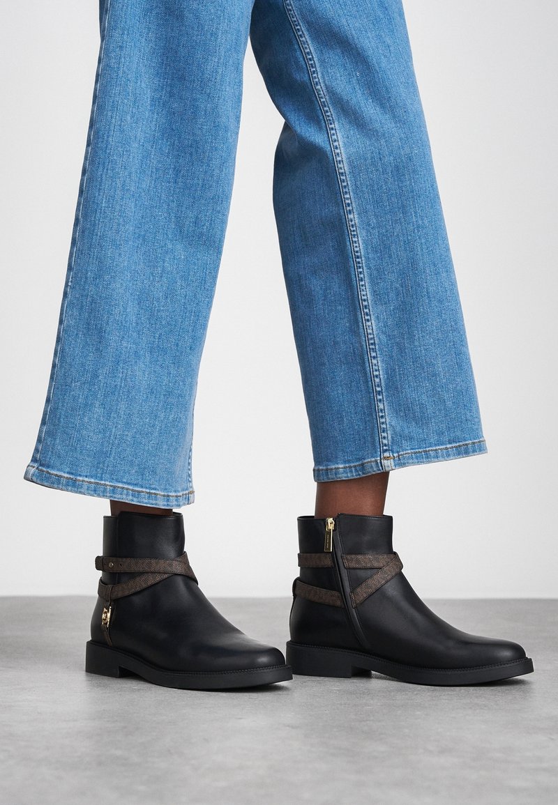 Personne portant un jean bleu à jambes larges et des bottines noires avec des sangles marron et des fermetures éclair latérales sur un sol gris.