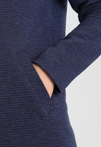 Mano parzialmente all'interno della tasca laterale di un indumento a maniche lunghe di un blu navy texture su uno sfondo chiaro.