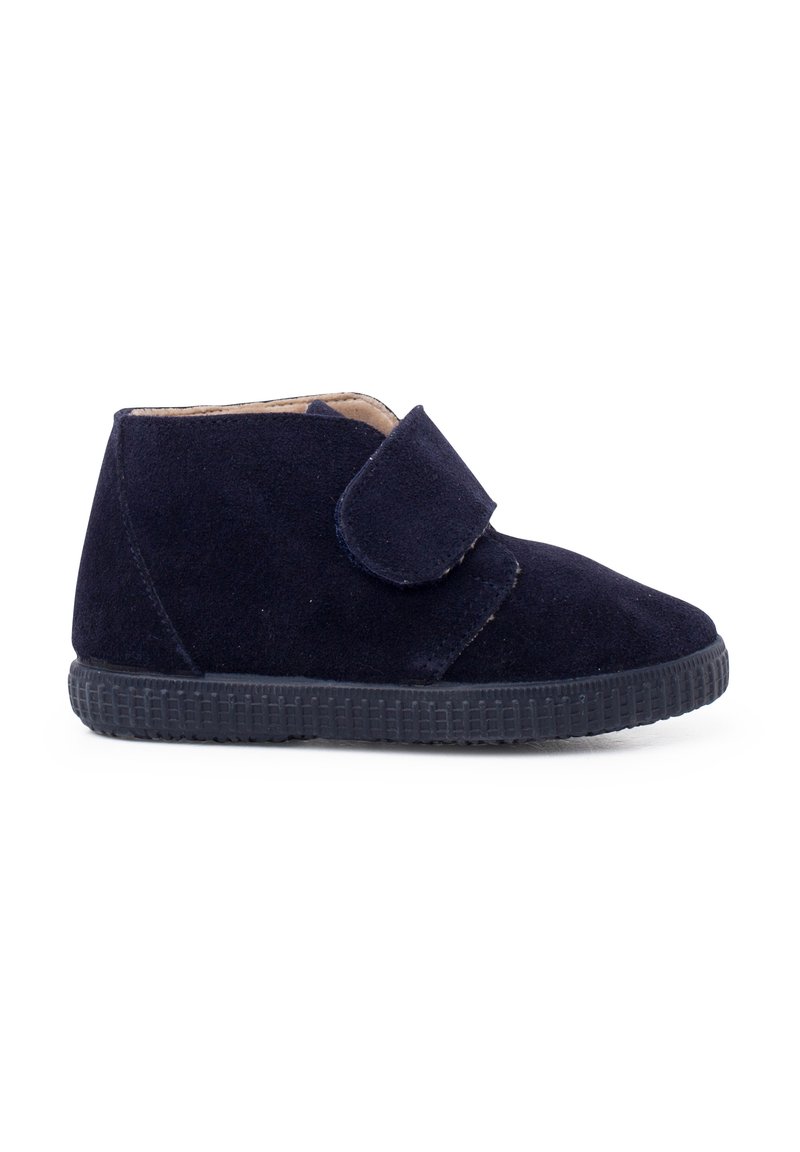 Zapato de ante azul marino para niños con tira de velcro y suela de goma negra texturizada, mostrado de perfil lateral sobre un fondo blanco.