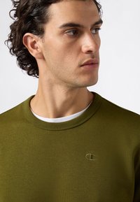 Oliwkowy sweter z okrągłym dekoltem, teksturowana bawełniana tkanina i małe haftowane logo na piersi. Widoczna biała koszulka pod spodem.