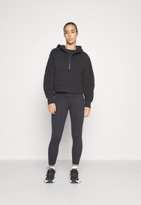Schwarze, kurze Kapuzenjacke mit halbem Reißverschluss, Raglanärmeln und Fronttasche, kombiniert mit schwarzen Leggings und schwarzen Turnschuhen. Glatte Stoffoberfläche.