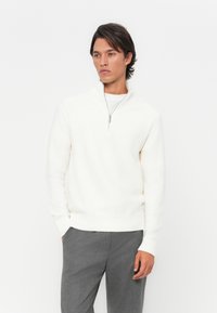 White zip-up sweater with ribbed cuffs and hem, smooth texture, worn over a white shirt, paired with gray trousers.