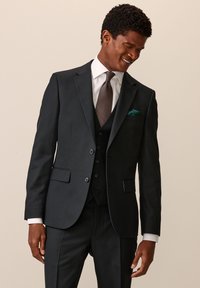 Abito nero su misura con un gilet a tre bottoni, camicia bianca e cravatta marrone testurizzata. Presenta un fazzoletto da taschino turchese e rever in raso.