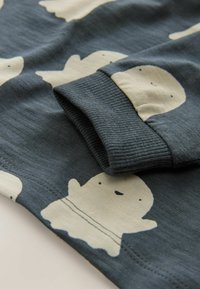 Blaugrauer Stoff mit einem sich wiederholenden Muster von lächelnden Gespensterfiguren in Creme. Gerippte Bündchen betonen die Ärmel. Weiche Textur.