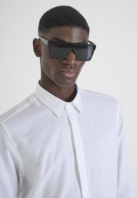 Occhiali da sole neri oversize con lenti piatte, design angolare; indossati con una camicia bianca, testurizzata, con bottoni e colletto classico.