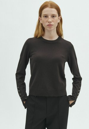 Jeune femme aux cheveux roux portant un pull noir à manches longues avec des boutons aux poignets et un pantalon noir, debout les mains dans les poches.