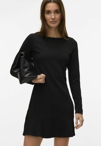 Vestido negro de manga larga con escote redondo y bajo acampanado, combinado con un bolso negro de textura suave. Material liso y ajustado.