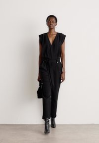 Mono negro con mangas cortas, escote en V, cintura ceñida con cinturón, parte delantera con pliegues, combinado con botas negras hasta el tobillo y sosteniendo un bolso negro pequeño.