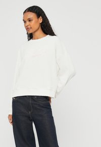 Sweatshirt oversized branco com bainha canelada e ombros caídos, com "HARBOUR INTERNATIONAL" bordado em rosa suave. Os jeans são azul-escuro.