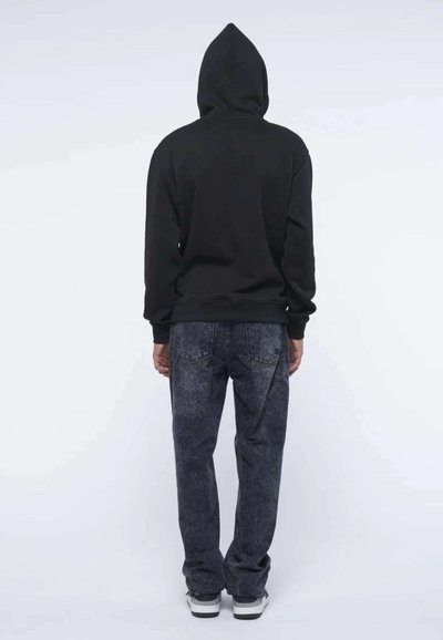 Felpa nera con cappuccio dallo stile rilassato, abbinata a jeans in denim scuro sbiadito. Tasche posteriori visibili e dettagli grigi sui jeans.