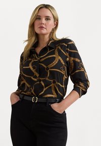 Svart knappskjorte med et gullkjede mønster, med langermede ermer rullet opp og spiss krage, kombinert med svarte jeans og et belte.
