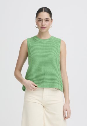 Žena s tmavými vlasmi v zelenom pletenom top bez rukávov a krémových nohaviciach pózujúca pred bielym jednoduchým pozadím.