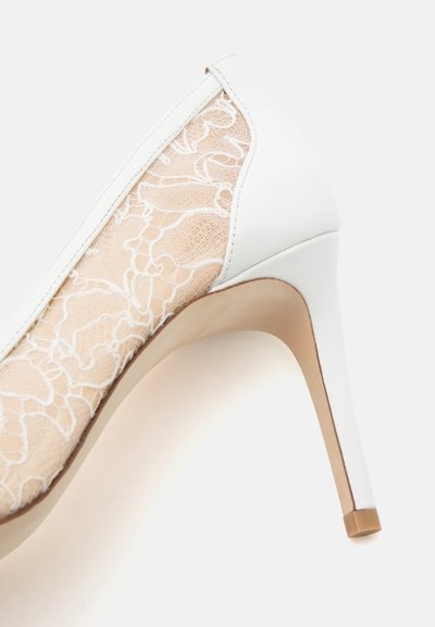Chaussure à talon haut blanche avec une superposition florale en dentelle beige et un talon en cuir lisse sur un fond blanc uni.
