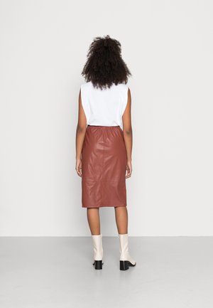 Femme aux cheveux bouclés portant un haut blanc sans manches, une jupe en cuir marron longueur genoux et des bottines crème à talons, de dos.