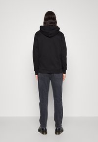 Persona con pelo oscuro a la altura de los hombros, vistiendo una sudadera negra, jeans gris oscuro y zapatos negros, de pie frente a una pared lisa de color claro.