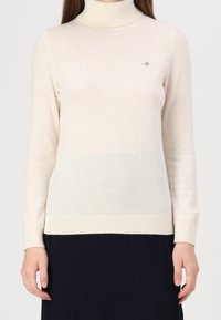 Béžový rolák z měkké pleteniny. Má dlouhé rukávy, žebrované manžety a malý emblém na hrudi. Bledne do světlejšího odstínu.