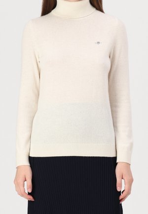Beige turtleneck sweater in soft knit fabric. Features long sleeves, ribbed cuffs, and a small emblem on the chest. Fades to a lighter shade.