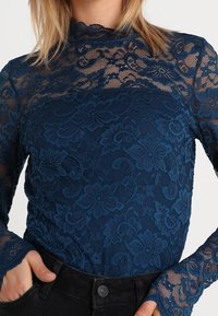 Maglietta in pizzo navy con collo alto e maniche lunghe, caratterizzata da motivi floreali e un design testurizzato. Abbinata a pantaloni neri aderenti.