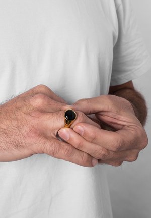 Bague en ton or avec une pierre noire ovale lisse. Le design présente un anneau effilé et une finition polie, tenue entre deux mains.