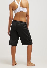 Donna che indossa un reggiseno sportivo bianco e pantaloni corti da basket neri larghi, in piedi a piedi nudi con la schiena rivolta verso la fotocamera.