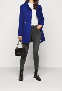 Blauer Mantel aus Wollmischung mit doppelreihiger Knopfleiste und schwarzen Knöpfen, kombiniert mit grauen Skinny-Jeans und schwarzen Stiefeletten mit Absatz. Schwarze Handtasche mit Kettenriemen.