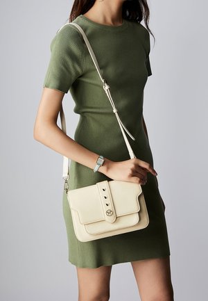 Kremowa skórzana torebka z klapką, trzema ozdobnymi ćwiekami i regulowanym paskiem, zestawiona z zieloną prążkowaną sukienką.