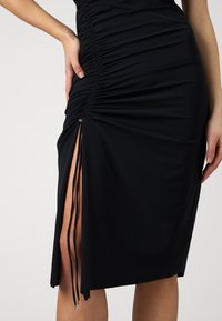 Sort kjole med ruched detaljering, en sideslitt med justerbare snorer, og en glatt, elastisk stofftekstur.