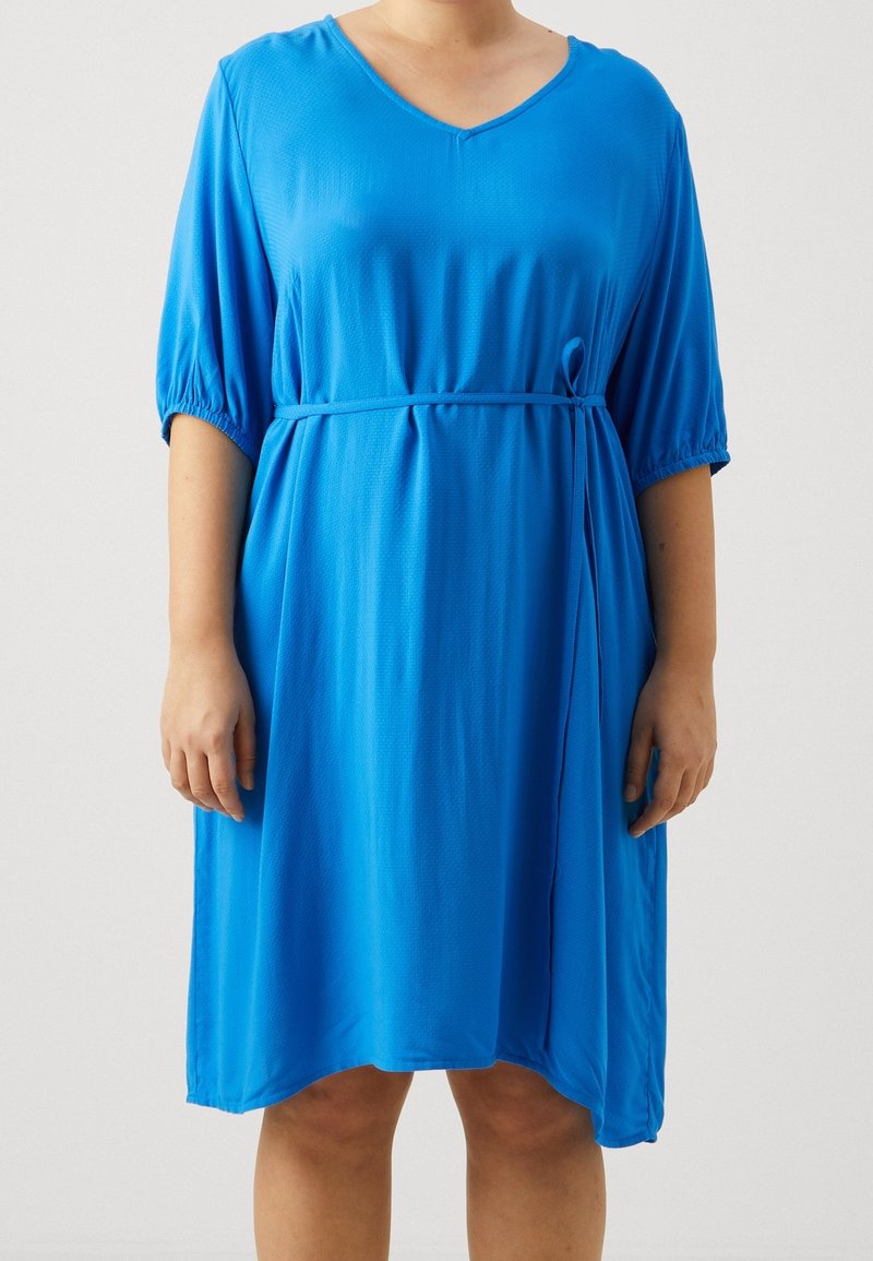 Femme portant une robe bleu vif arrivant au genou avec des manches bouffantes jusqu'au coude et une fine ceinture nouée à la taille.