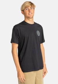 Black cotton t-shirt with short sleeves, crew neck, featuring a circular logo on the left chest and a relaxed fit. Worn with light corduroy pants.