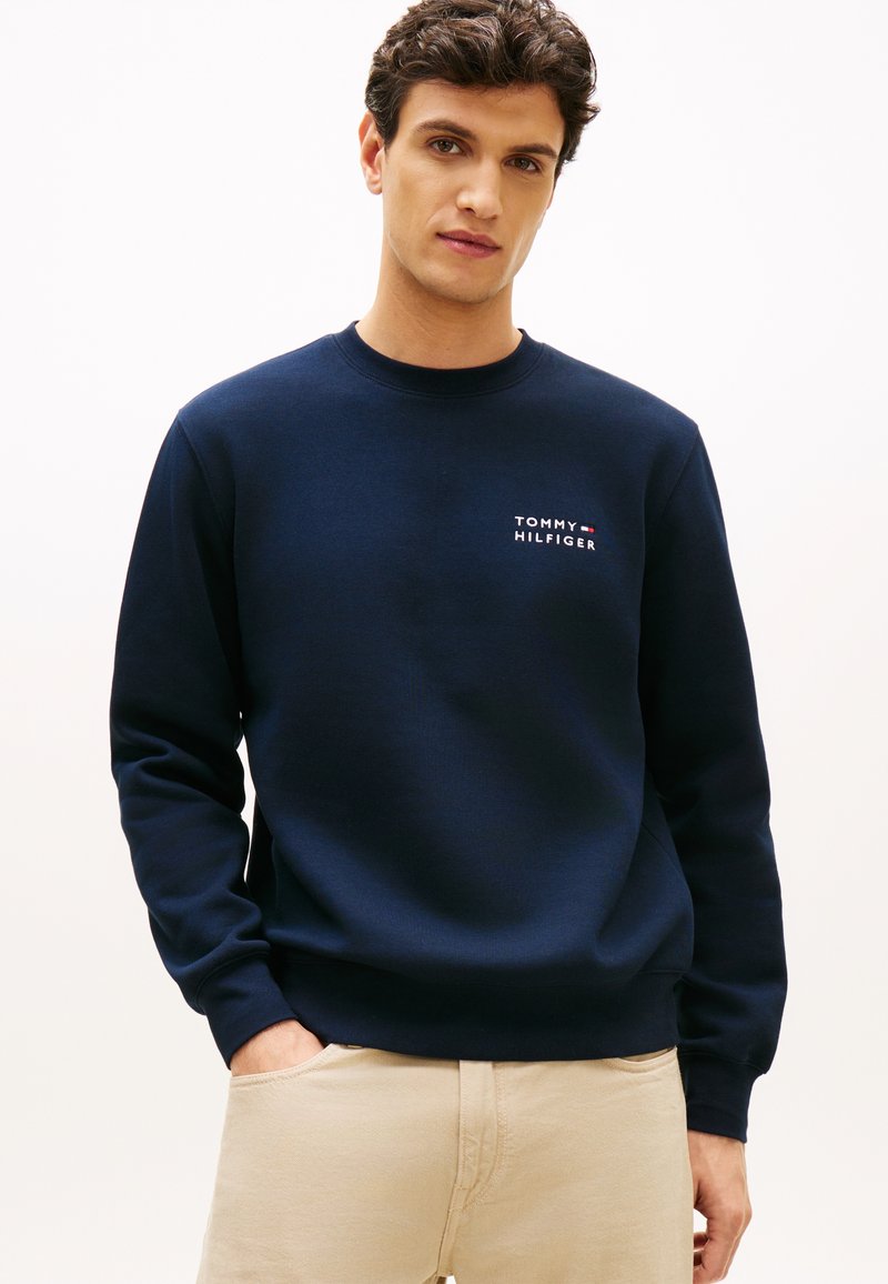 Νέος άνδρας που φοράει μπλε σκούρο φούτερ Tommy Hilfiger και μπεζ παντελόνι, στέκεται με το ένα χέρι στην τσέπη μπροστά σε λευκό φόντο.