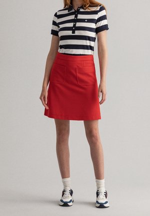 Czerwona spódnica z dwoma przednimi kieszeniami, w połączeniu z granatowo-białą koszulką polo w paski. Modelka nosi białe i niebieskie trampki z załogowymi skarpetkami.