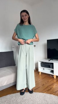 Ohlapna mint zelena majica z mehkimi plisiranji, v kombinaciji z visokim pasom in širokimi svetlo zelenimi hlačami. Črna ravna obuvala. Ozadje z leseno talno oblogo.