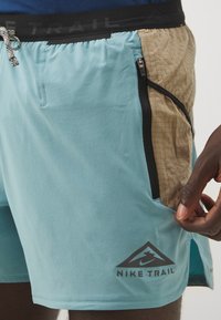 Gaiši zili sporta šorti ar strukturētu brūnu paneļu, melnu rāvējslēdzēju kabatu un Nike Trail logotipu uz kājas. Iekļauta aukliņa jostasvietā.